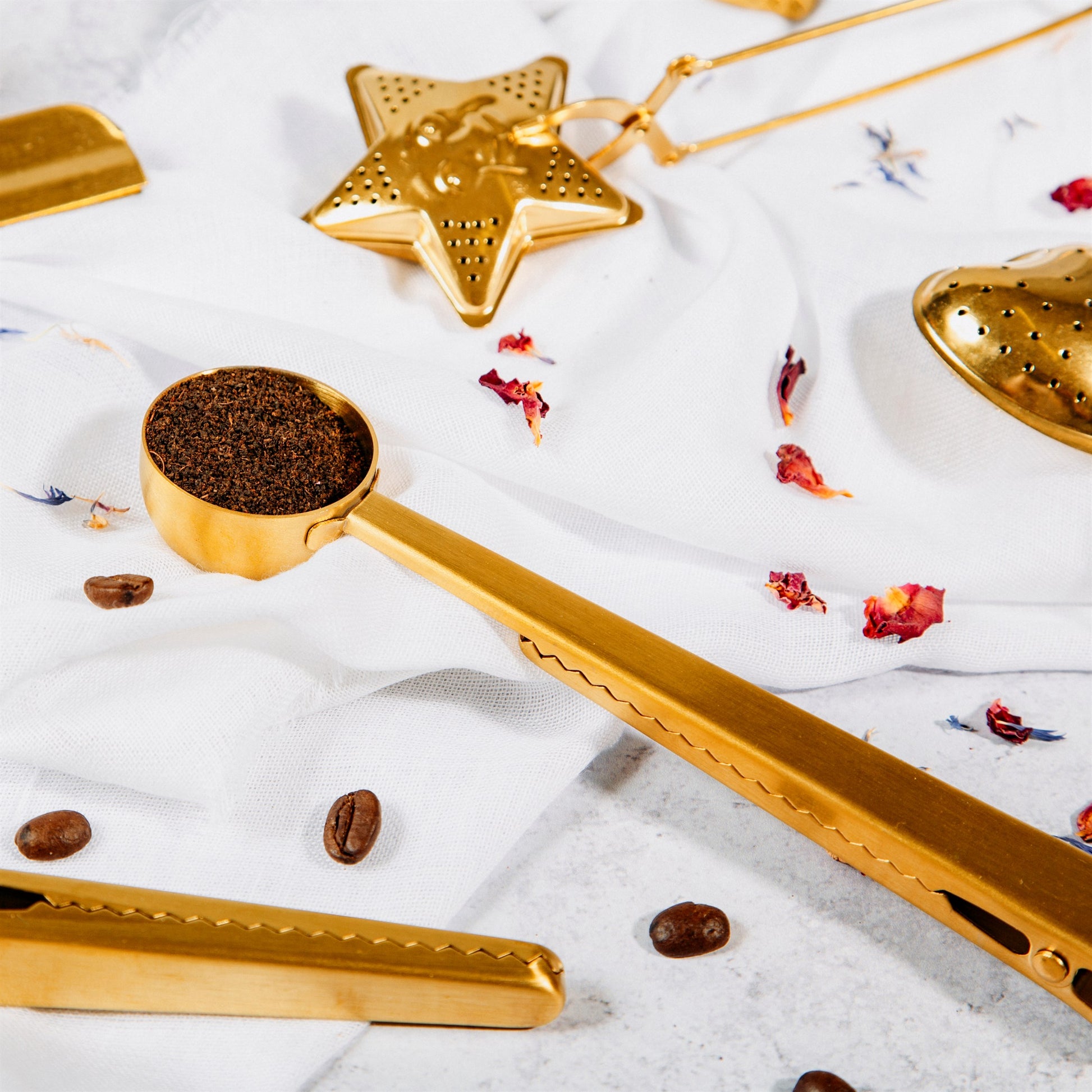 A gold stainless steel measuring scoop with an inbuilt clip, alongside scattered coffee beans and a star-shaped object, all placed on a white surface.