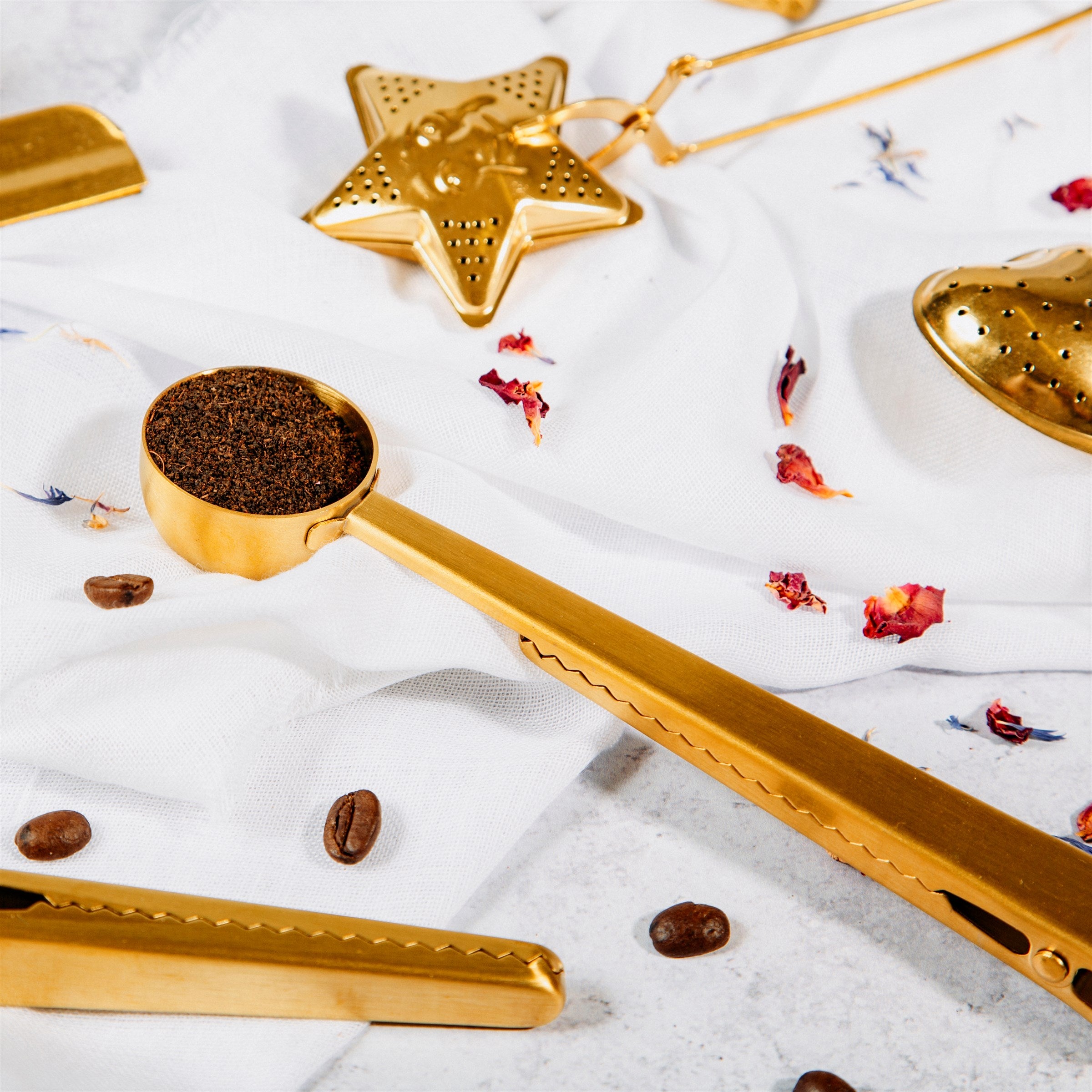 A gold stainless steel measuring scoop with an inbuilt clip, alongside scattered coffee beans and a star-shaped object, all placed on a white surface.